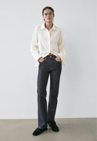 Camicia bianca con bottoni e colletto, abbinata a jeans scuri in denim e mocassini neri, in piedi davanti a uno sfondo neutro.