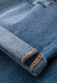Jeansmaterial i olika nyanser av blått, med en uppvikt manschett med brun kontrasterande söm och fransiga kanter, som visar textur och hantverk.