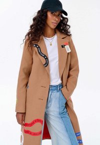 Žena s dlouhými kudrnatými vlasy má na sobě černou čepici, béžový kabát s barevnými nášivkami s nápisem "NEPŘESTÁVEJ", bílý tričko a světle modré džíny, pózující s rukama v kapsách.