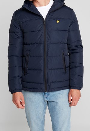 Mężczyzna ubrany w granatową pikowaną kurtkę z kapturem i żółtym logo ptaka, w połączeniu z jasnoniebieskimi dżinsami i białą koszulką.