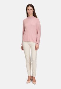 Helles rosa Strickpullover mit hohem Kragen und strukturiertem diagonalen Muster, kombiniert mit engen weißen Hosen und hellen Absätzen.