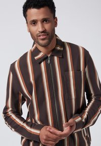 Brun och vit randig zip-up jacka med krage, framficka och långa ärmar. Tillverkad av ett strukturerat tyg.