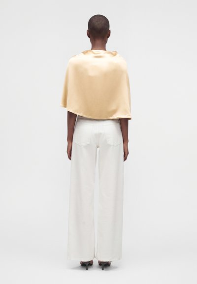 Personne aux cheveux courts portant une cape en satin beige, un pantalon blanc large et des talons noirs, debout de dos sur un fond uni.