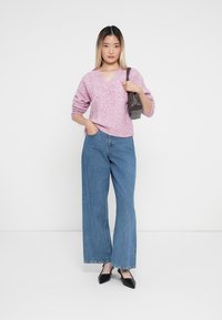 Rosa gestrickter Pullover mit V-Ausschnitt, kombiniert mit hochtaillierten, blauen, weit geschnittenen Jeans. Schwarze spitze Pumps und eine kleine graue Handtasche runden den Look ab.