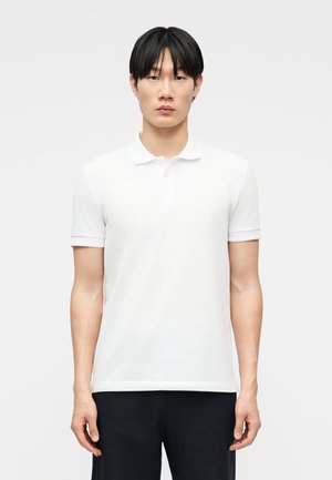 Jeune homme aux cheveux noirs et raides, portant un polo blanc à manches courtes uni et un pantalon noir, debout devant un fond clair.