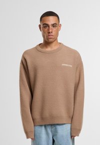 Hnědý pletený svetr s kulatým výstřihem a dlouhými rukávy. Má jemnou strukturu a malou bílou značku na přední straně.