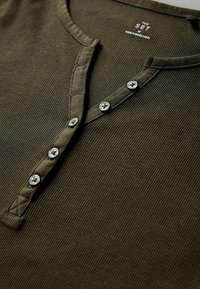 Oliven grønt ribbet henley-shirt med knaplukning, udstyret med fire grå knapper og en rund halsudskæring. Teksturdesign i stoffet.