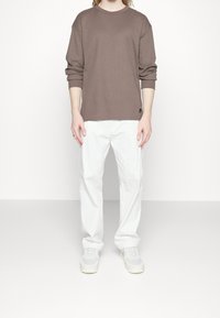 Hnědá košile s dlouhým rukávem a kulatým výstřihem, kombinovaná s bílými kalhotami a světlými teniskami. Materiál působí měkce; základní design bez vzorů.