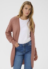 Femme avec de longs cheveux blonds portant des boucles d'oreilles créoles, un cardigan texturé rose poussiéreux, une chemise blanche et un jean bleu, les mains dans les poches.