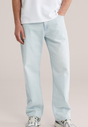 Férfi világoskék, egyenes szárú farmernadrágban, fehér és szürke sportcipőben, egyik kezével a zsebében áll.