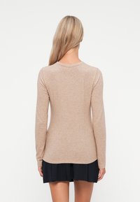 Beżowa koszula z długim rękawem i okrągłym dekoltem, o miękkiej fakturze, z lekko wygiętym dołem, noszona z ciemną plisowaną spódnicą.