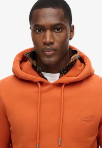 Sudadera naranja hecha de material suave con capucha ajustable, que presenta un logo en el pecho y una camisa de cuadros debajo.