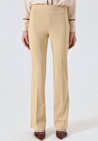 Beige Schlaghosen aus glattem Stoff mit hoher Taille. Sie zeichnen sich durch eine klare Silhouette aus und werden mit spitzen dunklen Schuhen kombiniert.