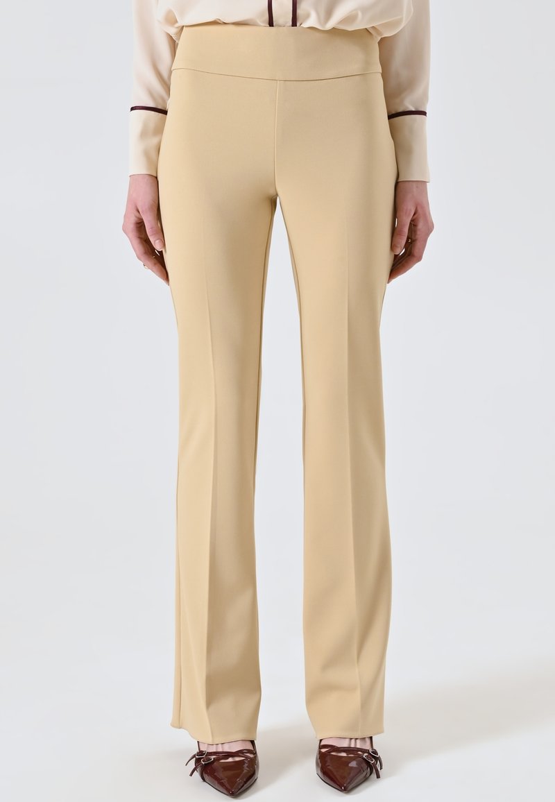 Beige Schlaghosen aus glattem Stoff mit hoher Taille. Sie zeichnen sich durch eine klare Silhouette aus und werden mit spitzen dunklen Schuhen kombiniert.