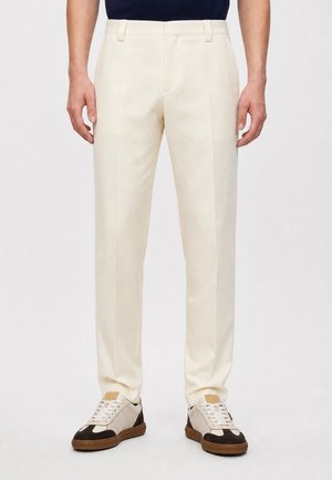 Man draagt crèmekleurige, op maat gemaakte broek met plooien aan de voorkant en witte en bruine casual vetersneakers.