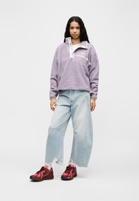 Νεαρή γυναίκα όρθια, φορώντας ένα λιλά φλις πουλόβερ, ανοιχτόχρωμα τζιν με φαρδιά ποδάρια και σκισίματα, λευκές κάλτσες και κόκκινα αθλητικά παπούτσια, πάνω σε λευκό φόντο.