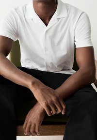 Weißes Poloshirt mit kurzen Ärmeln und einem breiten Kragen, das eine glatte Textur und eine Knopfverschlüsse aufweist. Modell sitzt mit verschränkten Armen.