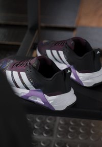 Ζευγάρι μωβ και μαύρα παπούτσια Adidas με λευκές ρίγες που στηρίζονται σε μαύρη επιφάνεια, δείχνοντας πλευρική και οπίσθια όψη.