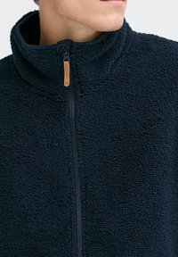 Marineblaue Fleecejacke mit hohem Kragen und Reißverschluss, ausgestattet mit einem Lederzieher und aus einem weichen, strukturierten Material gefertigt.