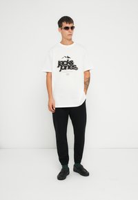 Camiseta blanca de manga corta con logo negro, combinada con pantalones negros ajustados y zapatos negros. El modelo lleva gafas de sol y calcetas verdes.