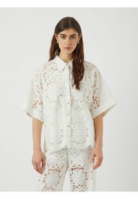 Vit spetsblus med blommönster, korta ärmar och knappstängning. Invecklade detaljer och lättviktsmaterial. Matchas med byxor.