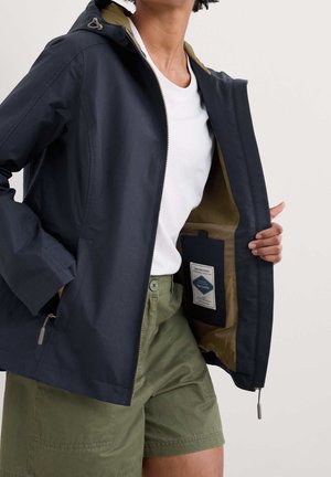 Person iført hvid skjorte, grønne shorts og navyjakke, der viser den indvendige etiket på den åbne jakke med lynlås og hætte.