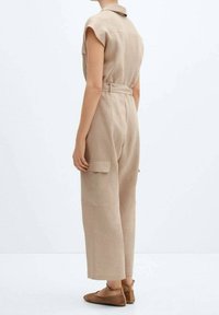 Combinaison en lin beige avec un col, des manches courtes, des poches latérales et une taille ceinturée ; portée avec des chaussures plates en tressé.