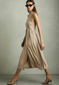 Beige midi ruha magas nyakkal, ujjatlan kialakítással és ráncolt szoknyával. Simább textúra és légies sziluett. Meztelen színű tűsarokkal párosítva.