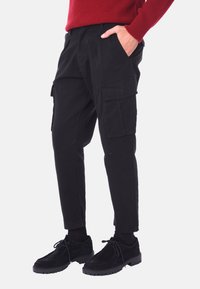 Pantaloni cargo neri in cotone con vestibilità slim, dotati di tasche laterali e una texture liscia. Indossati con scarpe nere.
