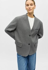 Blazer en laine grise avec un col en V, deux poches avant, et une fermeture à bouton unique. Porté sur une chemise blanche et accompagné d'un jean bleu.