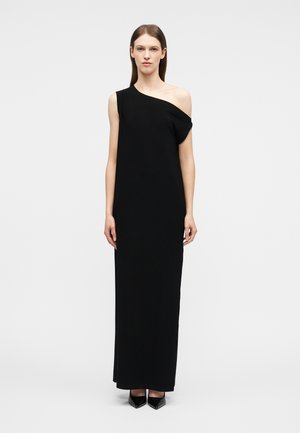 Donna che indossa un lungo abito nero monospalla con una manica drappeggiata, in piedi dritta contro uno sfondo grigio chiaro semplice.