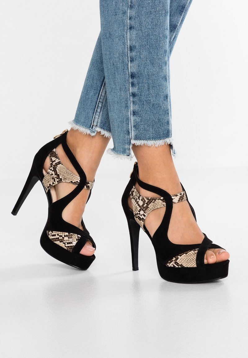 Femme portant un jean bleu à ourlet effiloché et des sandales à talons noirs avec un motif peau de serpent beige, debout sur un sol blanc.