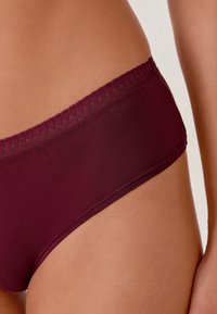 Slip in tessuto bordeaux con una superficie liscia, caratterizzato da un elastico con un motivo sottile. Il design è a vita bassa e aderente.
