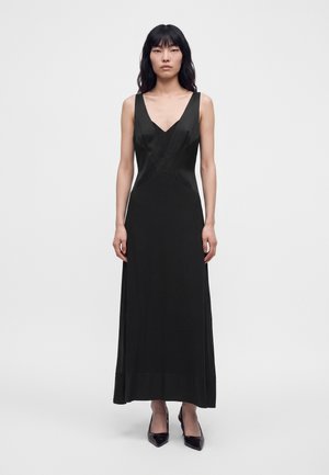 Une femme porte une longue robe noire sans manches à encolure en V et des escarpins à bouts pointus, sur un fond clair et uni.
