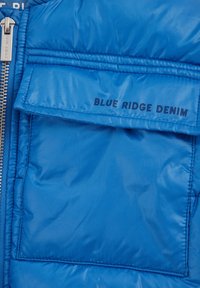 Kék puff лекокé fokozatú kabát, fényes felülettel, nagy első zsebbel, amelyen sötét nyomással a "BLUE RIDGE DENIM" felirat található, valamint fémből készült cipzárral.