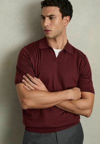 Polo din tricot de culoare bordo, cu mâneci scurte, guler și o croială lejeră. Tissue texturat, asortat cu pantaloni gri. Design simplu și curat.