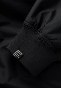 Poignet de veste en tissu noir avec bande élastiquée et étiquette tissée affichant du texte. Texture lisse avec un léger éclat. Design minimaliste.