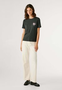 T-shirt nera a maniche corte con una grafica sulla parte superiore sinistra. Abbinata a jeans larghi di colore chiaro e scarpe nere.
