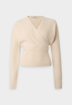 Femme debout, mains jointes, portant un pull côtelé crème, un jean large foncé et des ballerines noires sur un fond clair uni.