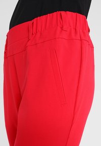 Pantalons rouges avec taille élastique, présentant une texture lisse, des poches latérales et un design slim. Le tissu semble léger et durable.