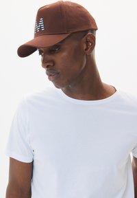 Brown baseball cap featuring an embroidered "M" in blue and white stripes, paired with a plain white t-shirt. Smooth fabric texture.