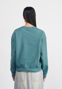 Tyrkysový oversized sveter s okrúhlym výstrihom, ribbed manžetami a spodným lemom. Zadná strana má hladkú textúru a žiadne viditeľné logá ani vzory.
