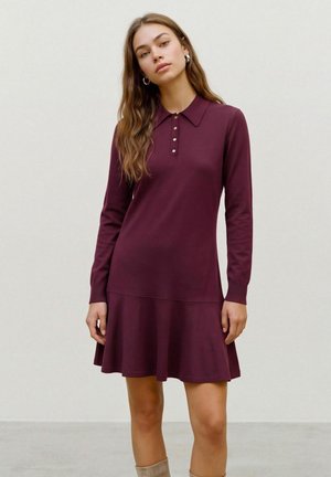 Une femme présente une robe bordeaux à manches longues, avec un col et une jupe évasée, sur un fond uni clair.