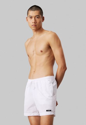 Białe szorty kąpielowe z elastycznym pasem i sznurkiem, z małym czarnym logo na dolnym brzegu, wykonane z lekkiego materiału.
