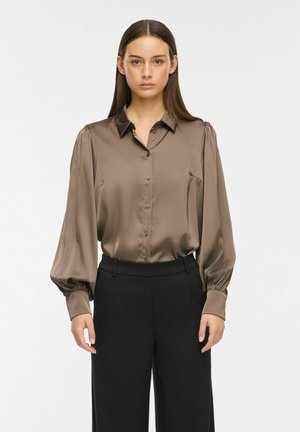 Femme aux cheveux raides et foncés portant un chemisier marron en satin à manches longues boutonné et un pantalon noir taille haute, debout sur un fond blanc.