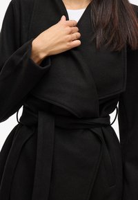 Abrigo negro envolvente hecho de una tela suave y texturizada. Presenta un cuello amplio, mangas largas y un lazo en la cintura a juego para una silueta ajustada.