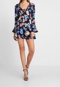 Romper dengan motif floral di latar belakang navy, lengan lonceng panjang, neckline v, dan detail ikatan di bagian depan. Sandal hak tinggi hitam dengan tali.