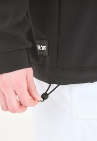 Veste de sport noire avec le logo SLOPE, dotée d'un cordon de serrage à l'ourlet et d'une texture lisse, présentée avec un pantalon blanc.