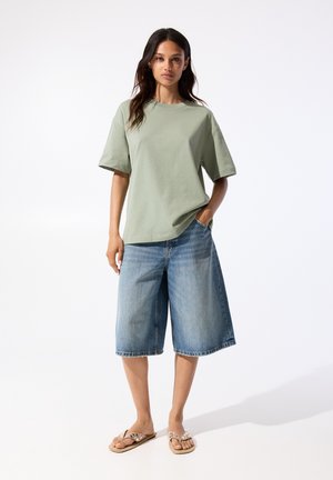 Hellgrünes übergroßes T-Shirt aus Baumwolle, kombiniert mit weiten blauen Denim-Shorts. Das Model trägt flache Sandalen und hat die Hände in den Taschen.