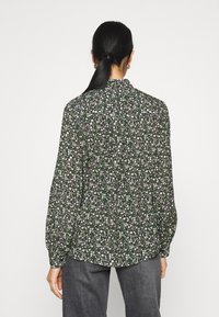Blusa a maniche lunghe in tessuto verde scuro, con un motivo floreale bianco, chiusura con bottoni sul retro e vestibilità ampia con polsini leggermente arricciati.